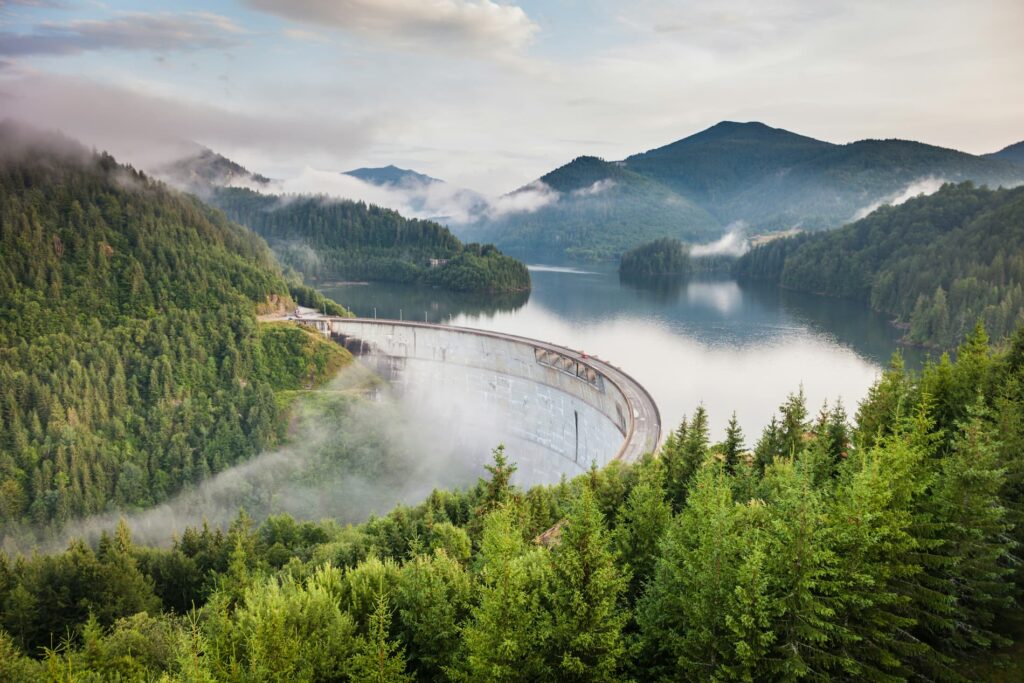 Une vue panoramique sur une vallée entourée de montagnes verdoyantes, avec un barrage au premier plan sous un ciel partiellement brumeux.
