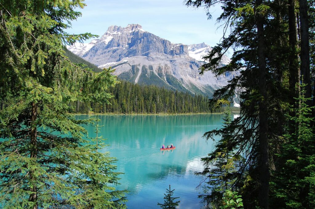 Lac turquoise entouré de montagnes enneigées et de forêts en Colombie-Britannique, Canada, avec un canoë rouge naviguant paisiblement sur l'eau.