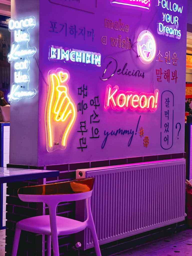 Un mur lumineux avec des néons colorés affichant des mots et expressions en anglais, coréen et des illustrations, créant une ambiance moderne et artistique liée à la culture coréenne