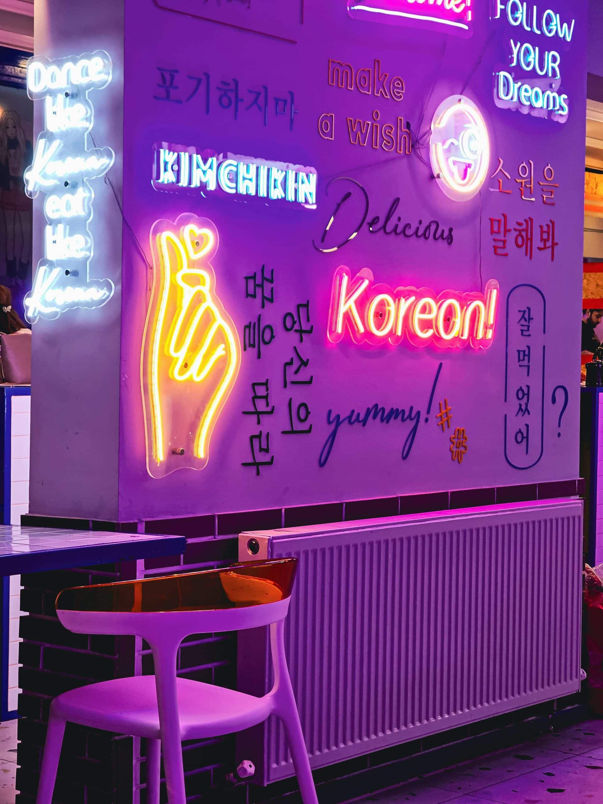 Un mur lumineux avec des néons colorés affichant des mots et expressions en anglais, coréen et des illustrations, créant une ambiance moderne et artistique liée à la culture coréenne