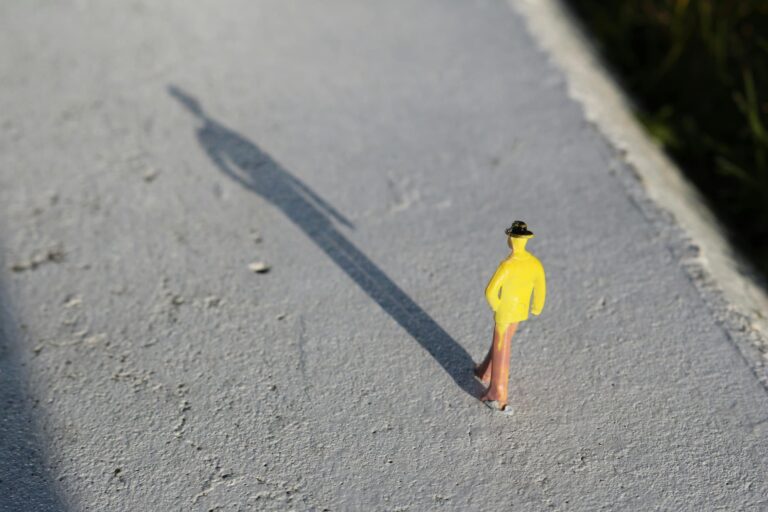 Une figurine en imperméable jaune marchant sur un chemin, projetant une longue ombre sur le sol. Image illustrant le concept de shadowing, une technique d’apprentissage des langues basée sur la répétition et l’imitation